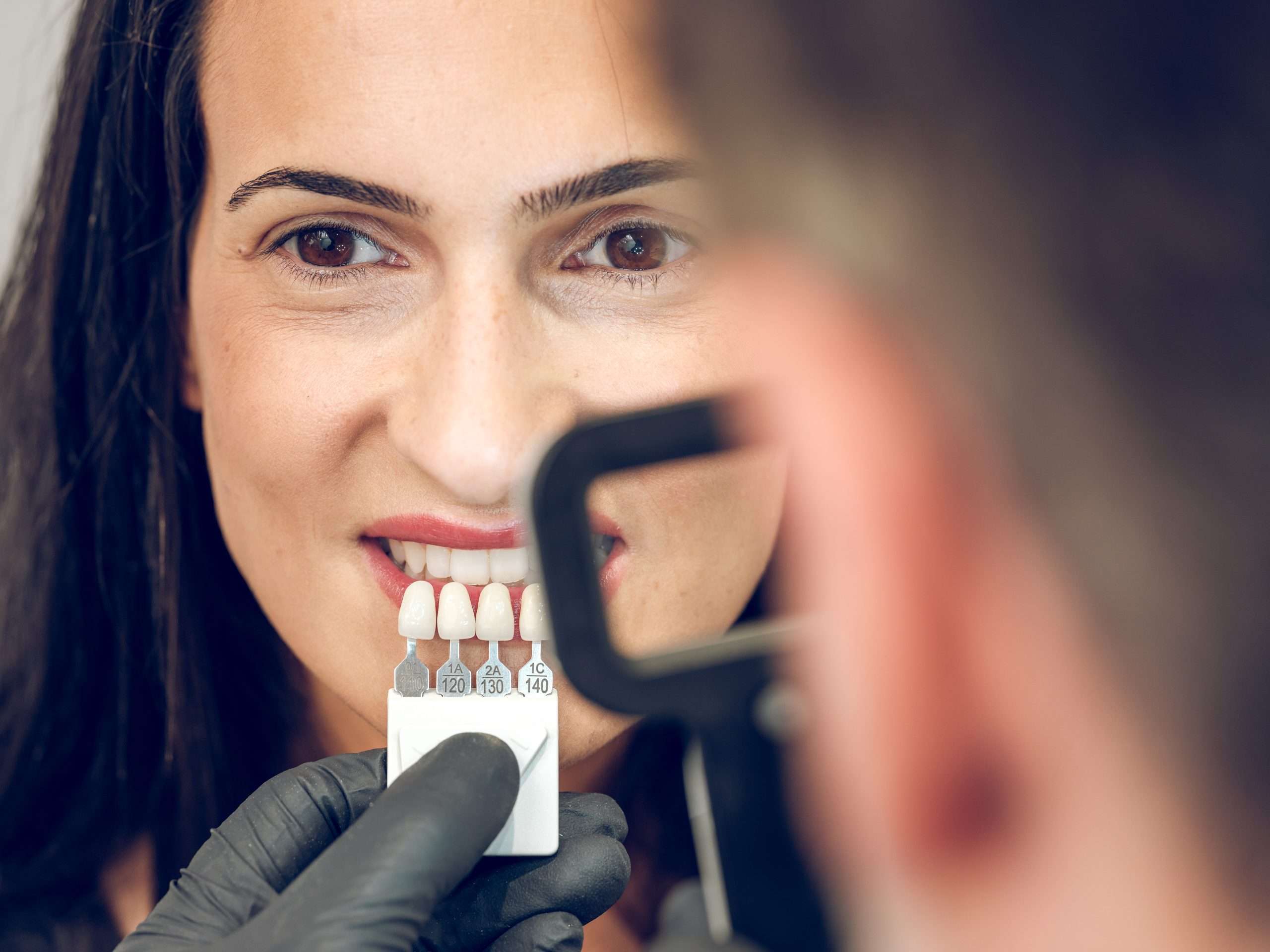 Dark haired lady looking at camera while crop blurred dentist selecting hue of teet